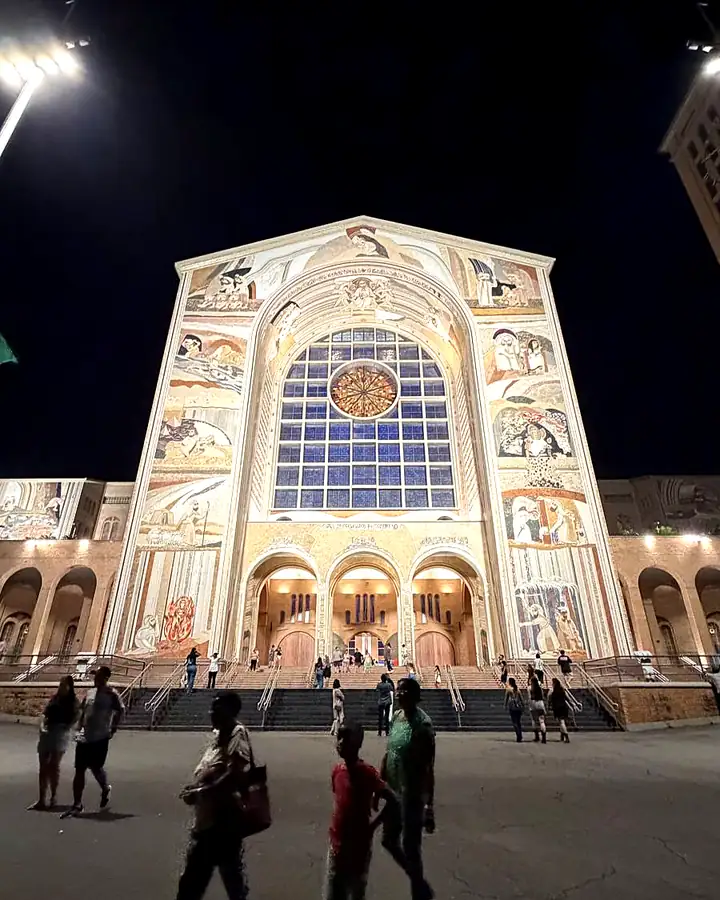 Fachada da Basílica de Aparecida à noite, com pessoas caminhando