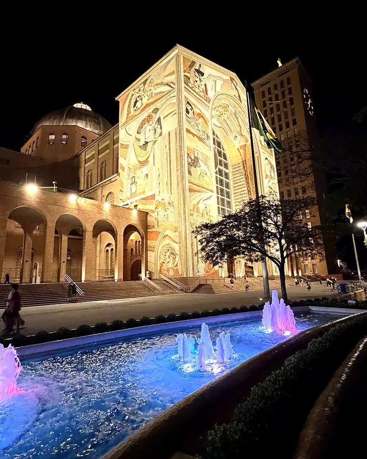 Basílica de Aparecida iluminada à noite, com o chafariz em primeiro plano